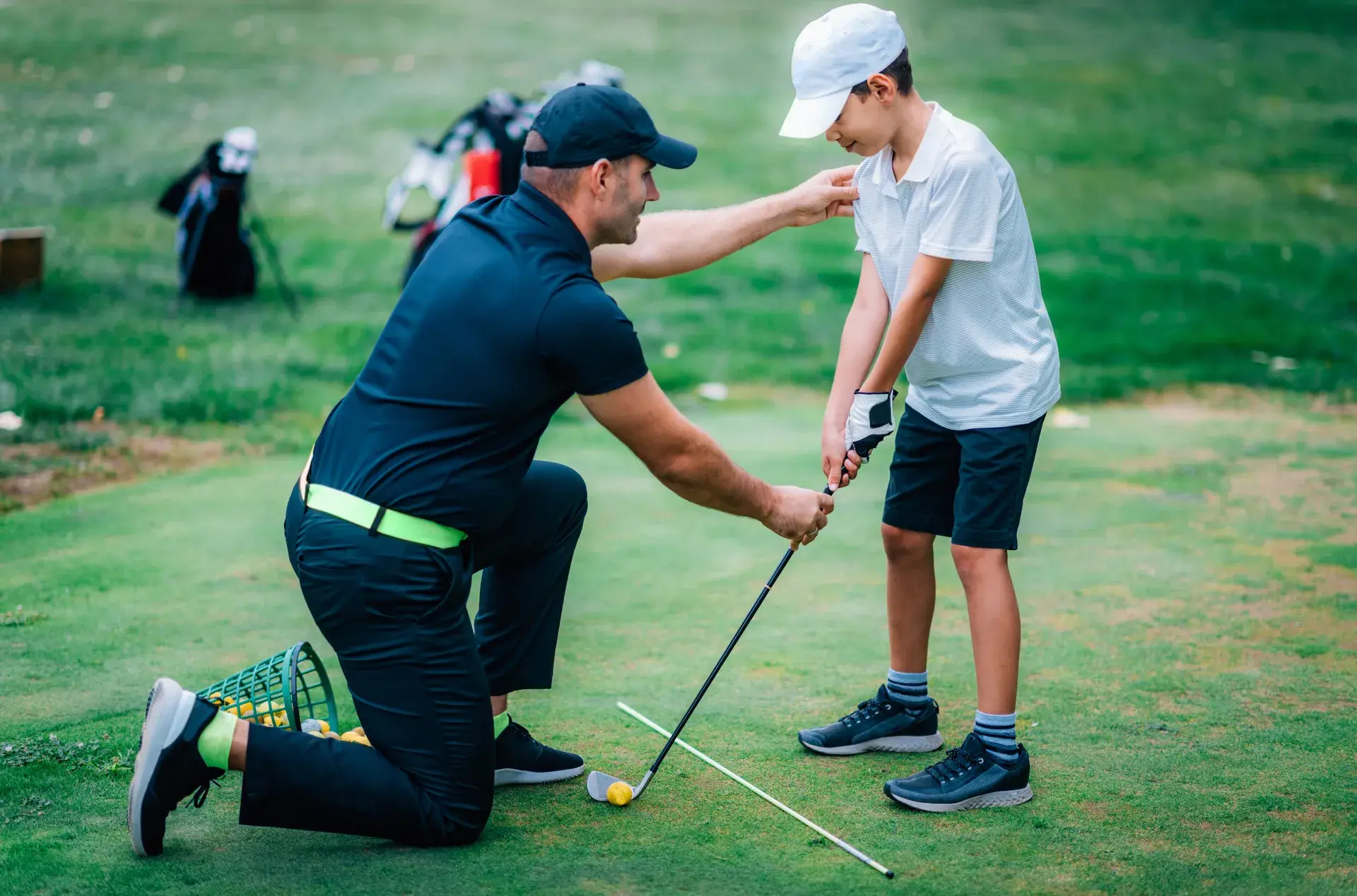 Coach kneeling beside a junior golfer, adjusting his grip and posture on the range